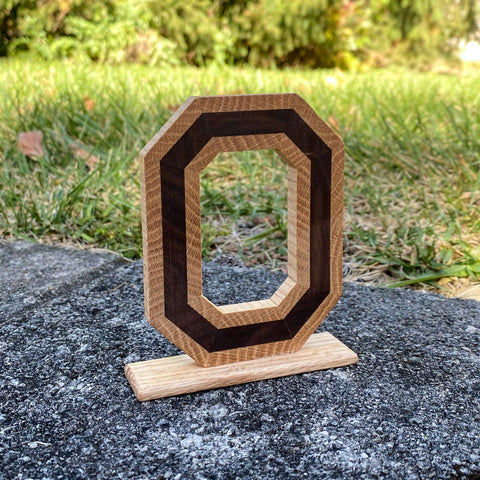 Wood crafted Block O letter from fallen walnut wood in the center and wrap of Oak wood around it, mounted on a Oak stand with a green grass background.